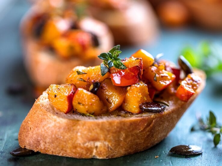 Eine Kürbis-Bruschetta mit Kürbiskernen und Kräutern auf einem Stück Baguette, im Hintergrund unscharf weitere Bruschettas. | © Adobe Stock/ karepa
