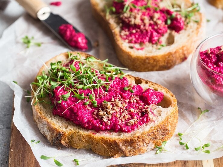 Ein paar Brotscheiben mit Pink Hummus, gehackten Nüssen und Kresse auf einem Holzbrett.