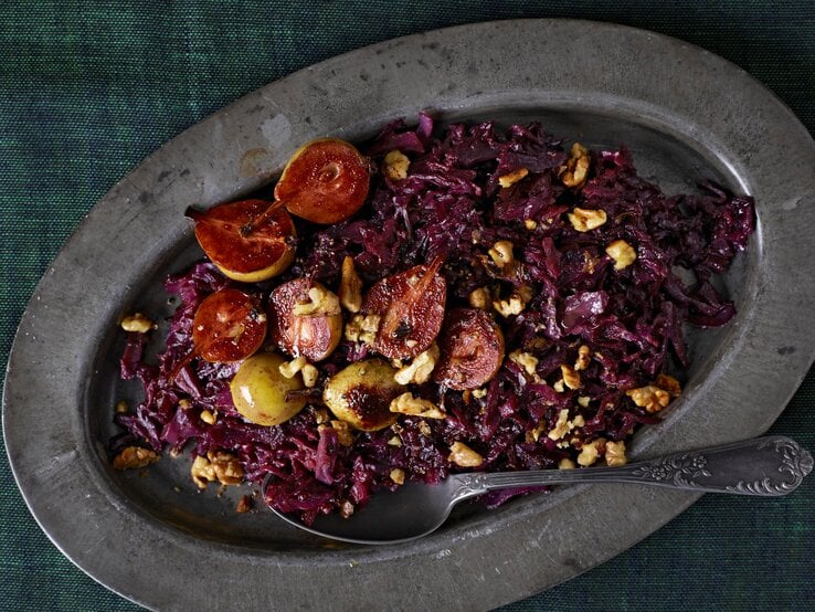 Eine ovale Platte auf der das geschmorte Rotkohl mit Birnen mit Walnüssen garniert und einem Löffel serviert wird, vor einem dunkelgrünen Hintergrund, von oben. 