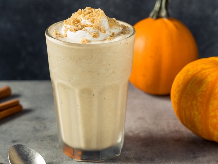 Frontalsicht: Ein Glas mit einem Kürbis-Milkshake mit Vanilleeis steht auf einem dunklen Untergrund. Neben dem Glas liegen Zimtstangen, eine Löffel sowie zwei kleine Kürbisse. | © Shutterstock / bhofack2