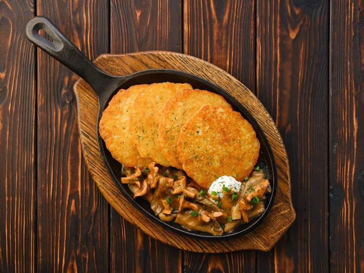 4 Kartoffelpuffer mit Rahm-Pfifferlingen in einer gusseisernen Pfanne, die auf einem Brett steht, Draufsicht.