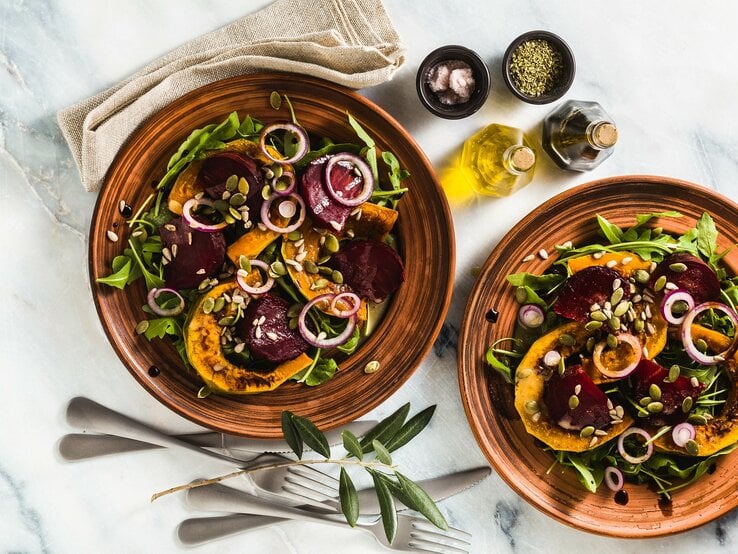 Zwei Teller Ofensalat mit Kürbis und Rote Bete sowie grünem Salat und Zwiebelringen, Draufsicht, daneben zwei Schalen mit Gewürzen und je eine Essig- und Ölflasche sowie eine Stoffserviette und Besteck.