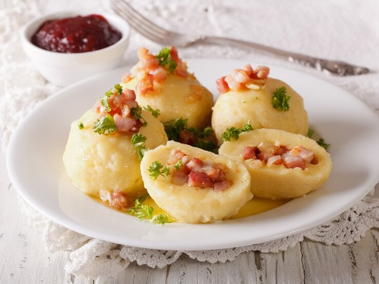 Frontal: auf einem weißen Untergrund aus Holz liegt eine weiße gehäckelte Decke auf der einen weißer Teller mit schwedischen Speck-Knödeln steht. In ihrer Heimat heißen die Kroppkator.