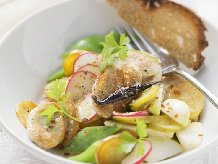 Ein weißer Tiefer Teller mit dem Bratwustsalat auf einem Tisch mit weißer Tischdecke sehr nah von schräg oben fotografiert.