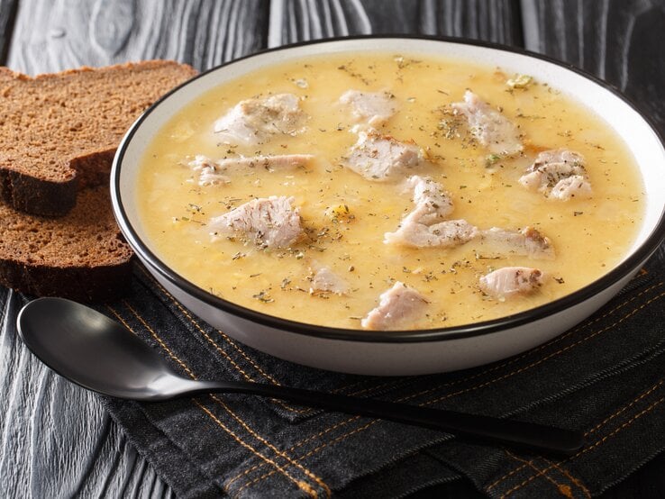 Ein voller Suppenteller mit der schwedischen Erbsensuppe mit Schweinefleisch, die Ärtsoppa neben einem Löffel und zwei Scheiben Schwarzbrot vor einem dunklen Hintergrund. 