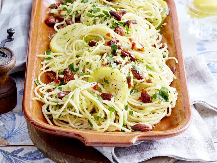 Zitronen-Mandel-Spaghetti, bestreut mit Chili und frischem Pfeffer sowie gehackter Petersilie, in einer Tonform auf gedecktem Tisch mit Tischdecke serviert. 
