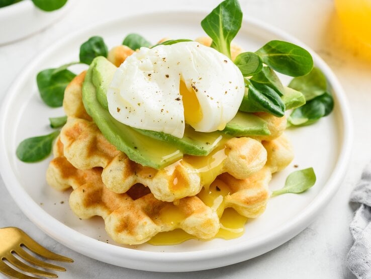 Käsewaffel mit Avocadoscheiben und pochiertem Ei oben, auf einem weißen Teller mit ein paar Feldsalatblättern garniert