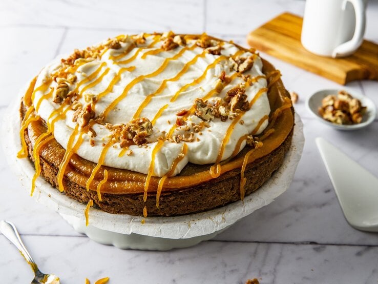 Kürbiskuchen mit Frischkäsecreme und gebrannten Walnüssen, garniert mit Karamell und serviert auf einer großen Kuchenplatte auf einem weißen Fliesentisch. 