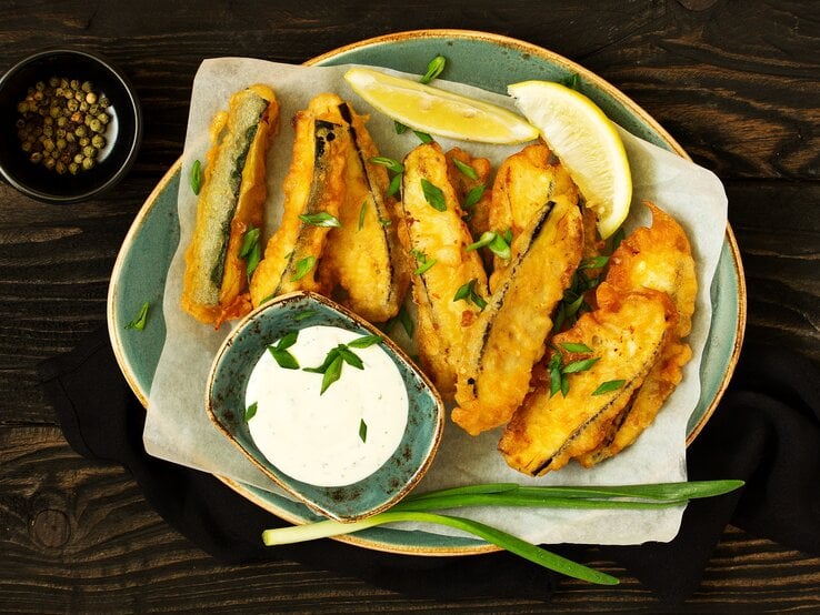 Draufsicht: asiatische Zucchini-Sticks die in einer Pfanne frittiert wurden. Daneben liegen zwei Zitronenschnitzer, eine Schale mit Pfefferkörnern und ein Schale mit Joghurt-Dip. | © Shutterstock / Lesya Dolyuk