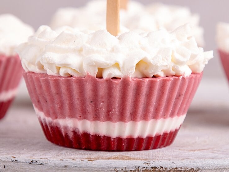 Ein Frozen-Yogurt-Muffin mit drei verschiedenen Schichten, mit Schlagsahne dekoriert, im Hintergrund unscharf und angeschnitten weitere Muffins.