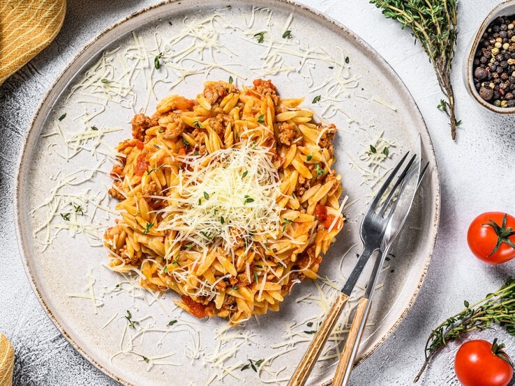 Ein Teller veganes Orzorisotto in der Draufsicht, mit veganem Parmesan bestreut, daneben Besteckt, ein Geschirrtuch, Tomaten und Gewürze.