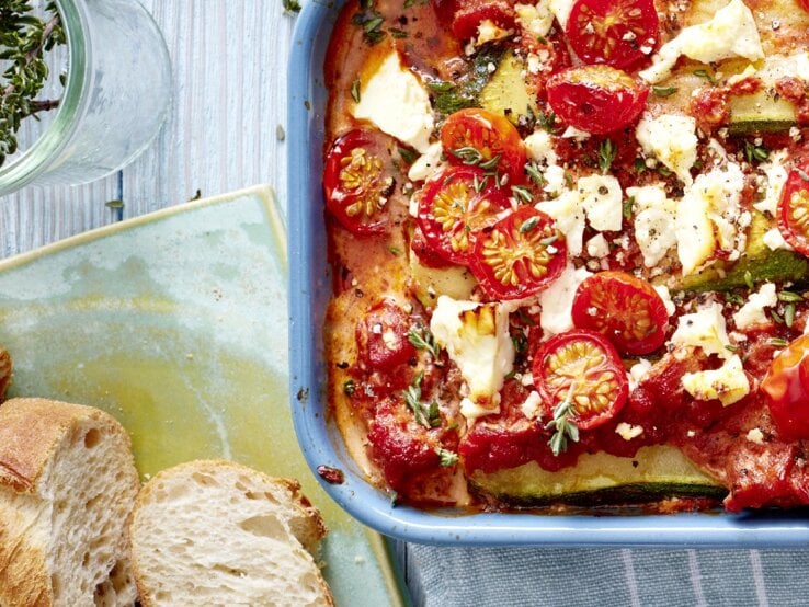 Eine Zucchini-Lasagne mit Feta in einer blauen Auflaufform steht auf einem Geschirrtuch auf einem blauen Tisch. Die Lasagne ist angerichtet mit Feta, Tomaten und Thymian. Neben der Auflaufform liegt Baguette und ein Glas mit Kräutern.