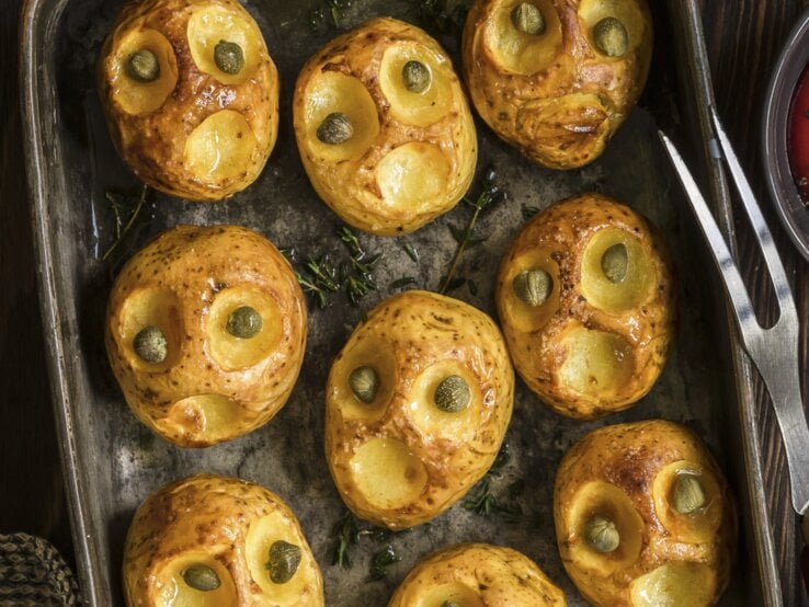 Halloween-Bratkartoffeln mit Kapernaugen, geschnitzt mit gruseligen Fratzen und serviert auf einem schwarzen Blech auf hölzernem Untergrund. 