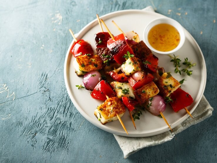 4 Tofu-Grillspieße mit Gemüse auf einem Teller, auf dem auch eine Schale mit Dip steht. 