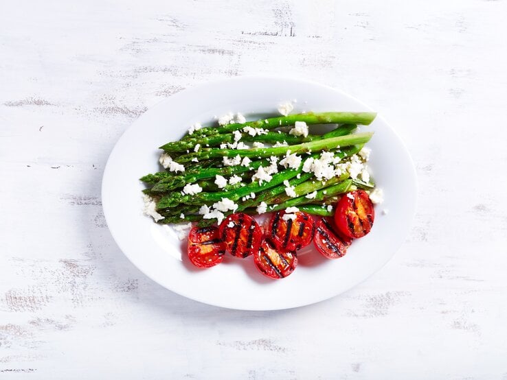 Ein Teller gegrillter Spargel mit Zitrone und Feta sowie gegrillten Tomaten in der Draufsicht.