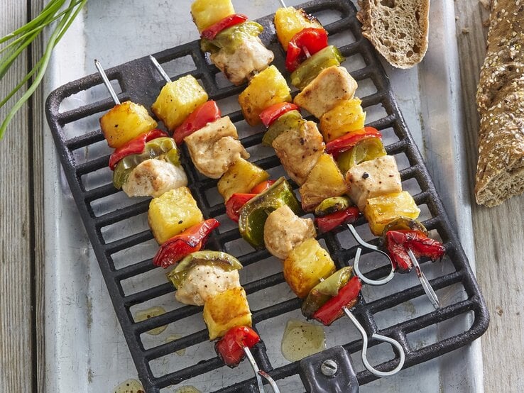 Mehrere Hawaii-Spieße mit Huhn und Ananas auf einem Grillgitter, daneben Brot in der Draufsicht.