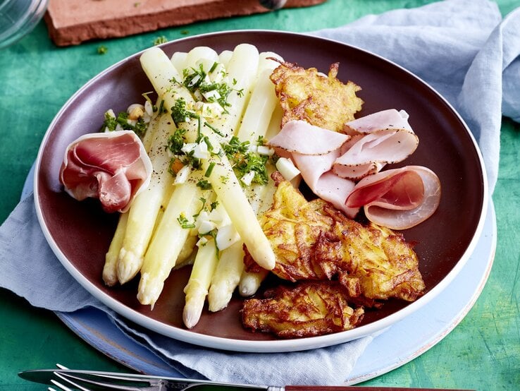 Auf einem Teller ist eine Portion Spargel mit Rösti und Schinken angerichtet. Sie ist mit Aufschnitt verfeinert.