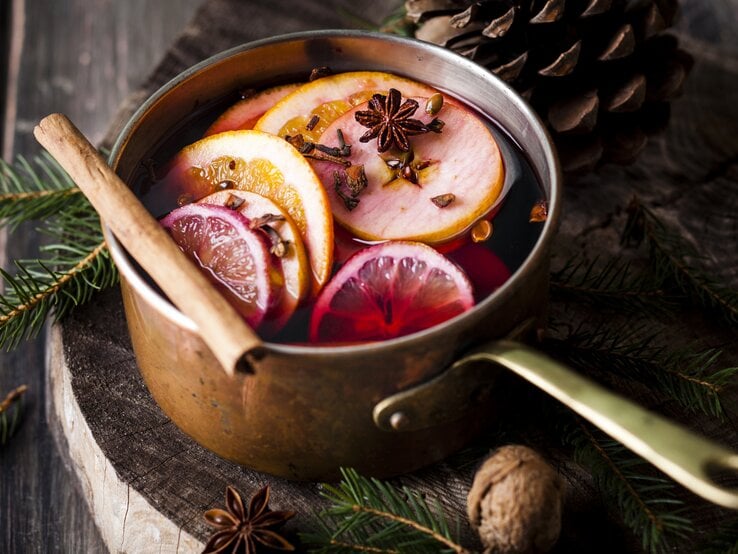 Ein Topf mit Glühwein, Orangenscheiben und Gewürzen steht auf einem Holzuntergrund, daneben liegen Tannengrün und Tannenzapfen.