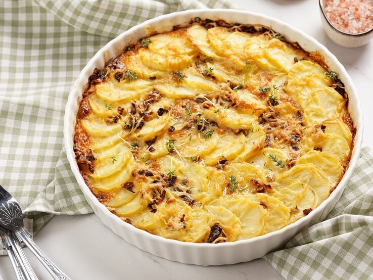 Eine runde weiße Auflaufform mit dem Kartoffelgratin auf einem weißen Tisch mit grün-weiß-karierter Tischdecke. | © Shutterstock/ RomanaMart 