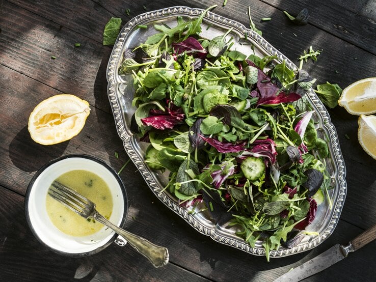 Silberne Platte mit Salat auf hölzernem Untergrund. Daneben einige ausgepresste Zitronen und eine Schale mit Buttermilch-Zitronen-Dressing. 