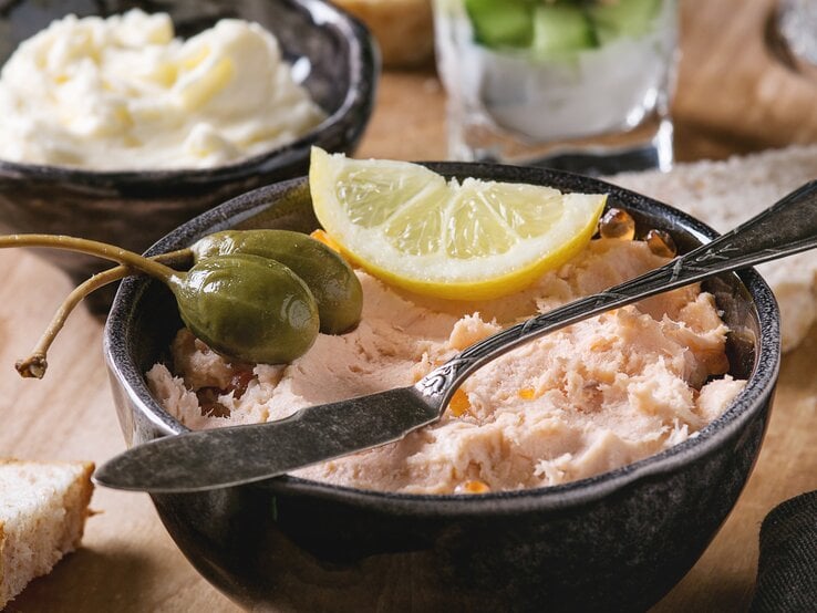 Frontalsicht: ein Räucherlachs-Dip mit Schmand in einemr schwarzen Schüssel. Darauf liegen Kapernäpfel und eine Zitronenscheibe. Im Hintergund stehen ein Schälchen Butter und ein Glas mit Räucherlachdip.