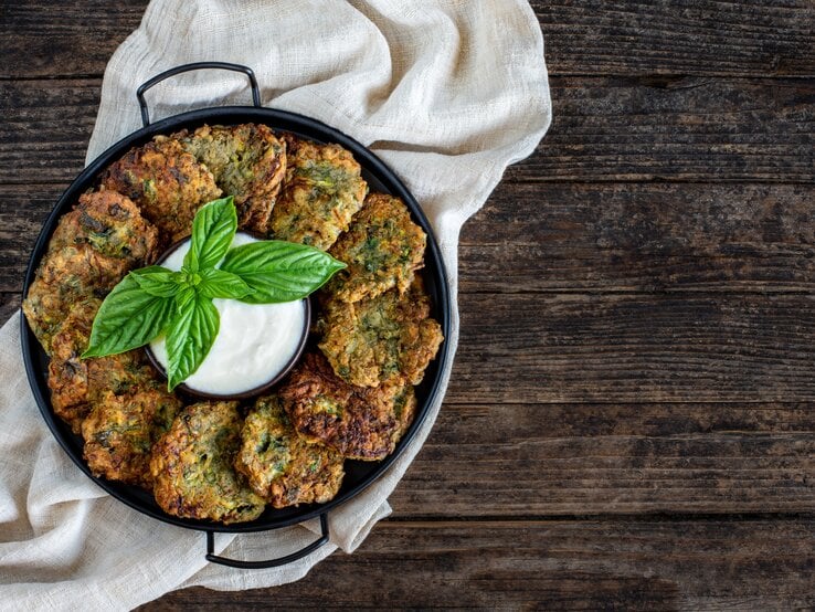 Eine Pfanne mit Brokkoli-Kartoffelpuffern, die im Kreis angeordnet sind, in der Mitte eine Schale mit Dip. Draufsicht.
