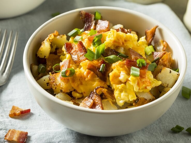 Eine weiße Schale mit dem Rührei auf grauem Untergrund mit Speck und Frühlingszwiebel. Alles von schräg oben fotografiert.