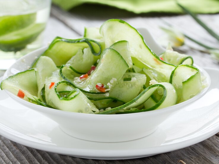 Eine Schale Gurkensalat mit Orangendressing und Kräutern auf einem weißen Holztisch.