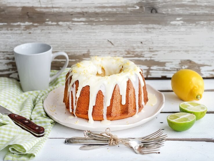 Ein Himbeer-Gugelhupf mit weißer Schokolade auf einem runden Teller, drumherum Limetten, Zitronen, eine Tasse, Kuchengabeln, ein Messer und ein Geschirrtuch.