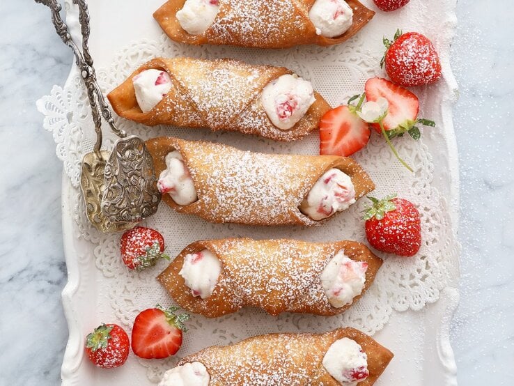 Italienische Cannoli mit Ricota-Erdbeer-Füllung, bestäubt mit Puderzucker und serviert auf einem weißen, verzierten Kuchenteller, angereicht mit frischen Erdbeeren.  | © StockFood / Jalag / Neubauer, Mathias