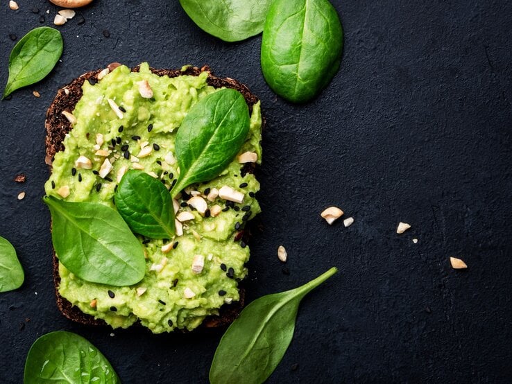 Draufsicht: Dunkler Untergund, ein Avocado-Cashew-Sandwich, dass mit schwarzen Sesamkörnern garniert ist. Drumherum liegen Spinatblätter.