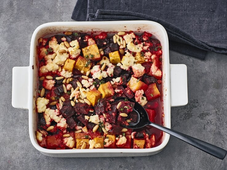 Rote-Bete Gratin mit Feta, Kürbis, Pinienkernen und Thymian in einer großen Auflaufform auf steinernem Untergrund. 