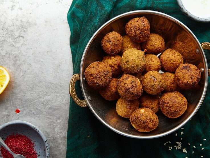 Eine silberne Schale gefüllt mit Maronen-Falafel mit Cranberrys neben einer Schüssel mit roten Gewürz, einer ausgepressten Zitronenhälfte und einer Schüssel mit einem Joghurt-Dip, vor einem dunkel grün und grauen Hintergrund, von oben. 