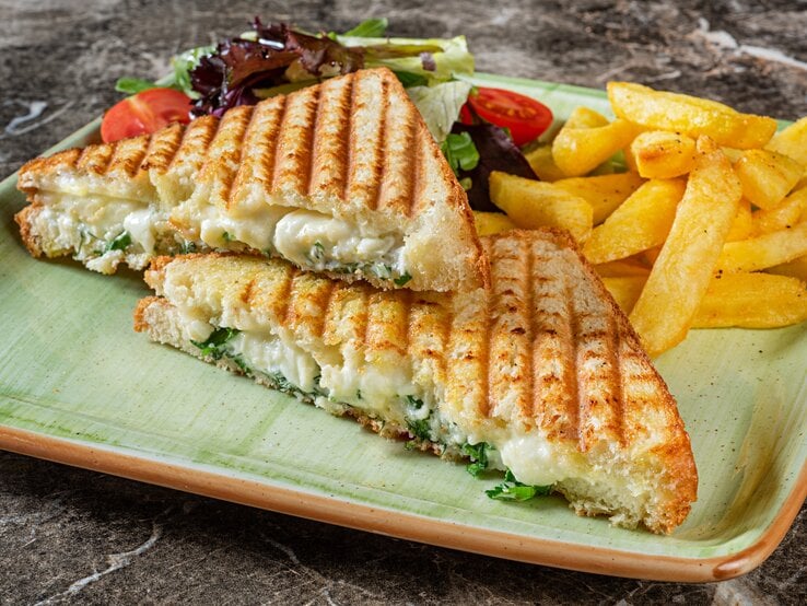 Auf einem eckigen Teller liegen zwei Panini mit Brokkoli und Käse gefüllt, außerdem Salat und Fritten.