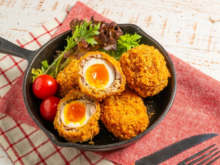 Eine Pfanne mit den schottischen Eiern von oben auf einem Tisch mit rot-weiß karierter Tischdecke fotografiert. | © Shutterstock / norikko