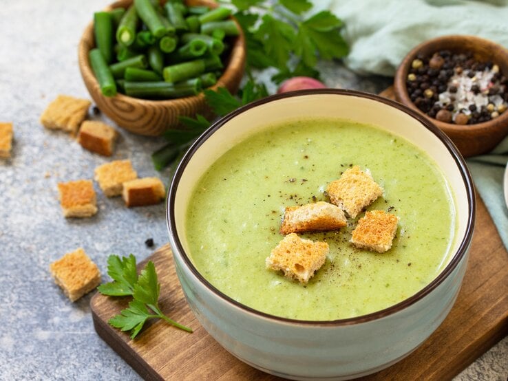 Draufsicht: Eine Schüssel mit einer grünen cremigen Flüssigkeit steht auf einem Holzbrettchen. Auf den Flüssigkeit liegen brotstücke. Neben der Schüssel liegen Petersilie sowie einige Gewürze.