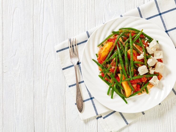 Aus der Vogelperspektive: Ein Teller Fasolakia lathera, einem griechischem Bohnentopf mit Kartoffeln, liegt auf einem karierten Tuch. Der Untergrund ist hell.  