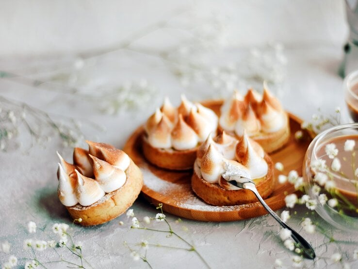 Vier Mini-Törtchen mit Wintercreme und Baiser aus Plätzchenteig auf Holzbrett. Daneben zwei Gläser mit Kaffee oder Kakao. Im Bild Blüten als Deko verteilt. Frontalaufnahme.