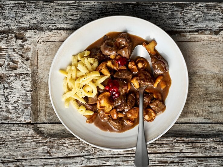 Ein weißer Teller mit einer Portion des Rehgulaschs mit Spätzle und Preiselbeeren, verfeinert mit Pfifferlingen. Ein Löffel ist auf dem Teller platziert, der vor einem Holzhintergrund von oben dargestellt ist.  