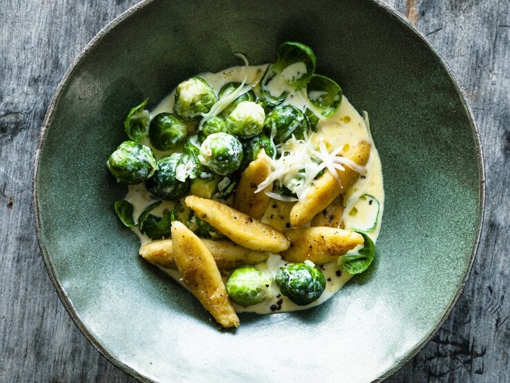 Schupfnudeln und Rosenkohl in heller Soße mit geriebenen Käse. Serviert in mattgrüner Schale auf grauem Holztisch.