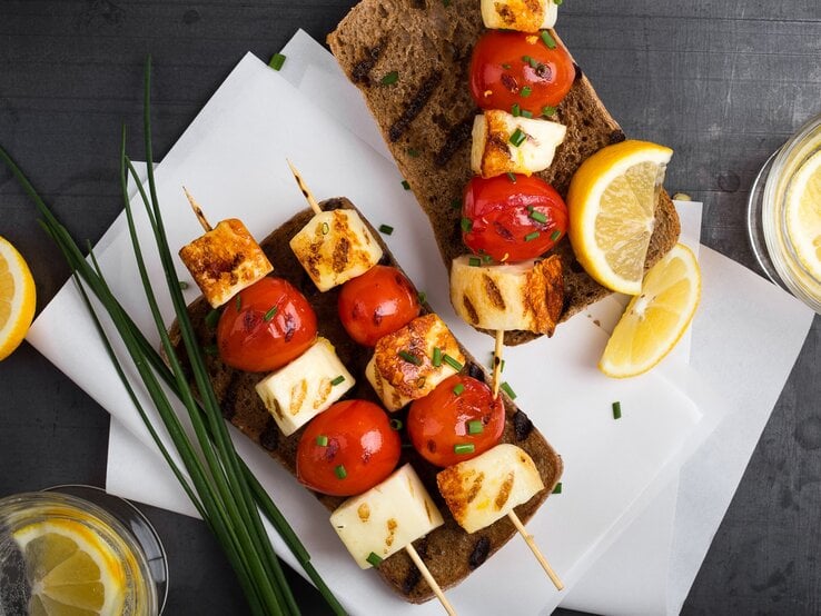 Halloumi-Tomaten-Spieße auf Brotscheiben. Daneben Zitrone und Schnittlauch. 
