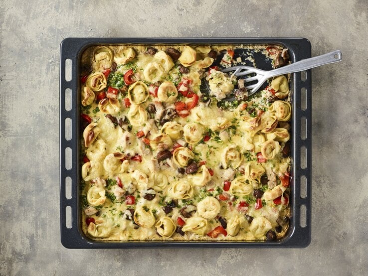 Überbackener Tortelloni-Auflauf auf Backblech mit knackiger Paprika, Kräutern, knusprigem Speck in cremiger Sahnesoße auf steinernem Untergrund