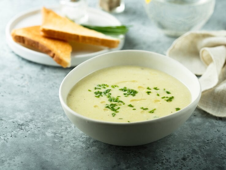 Eine weiße Schale mit der grünen Suppe auf bläulichem Untergrund. Alles von der Seite fotografiert.