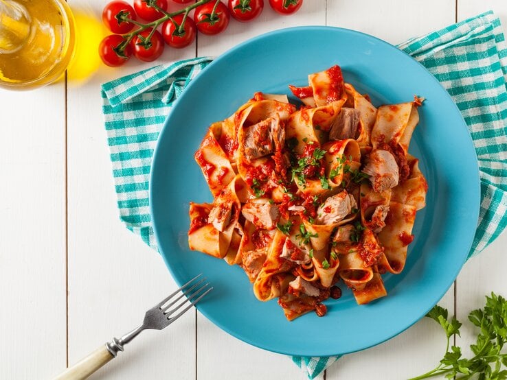 Ein Teller mit einer Portion One-Pot-Pasta mit Thunfisch und Tomatensoße, bestreut mit Petersilie. daneben liegen Tomaten, Petersilienzweige, außerdem ist eine Flasche mit Öl zu sehen.