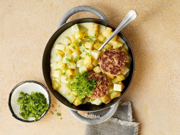 Eine tiefe Pfanne mit den Tatar-Frikadellen mit Kohlrabi-Kartoffel-Ragout, die mit Kerbel garniert sind, neben einer kleinen Schüssel mit Kerbel und einem Tuch, vor einem beigen Hintergrund, von oben. 