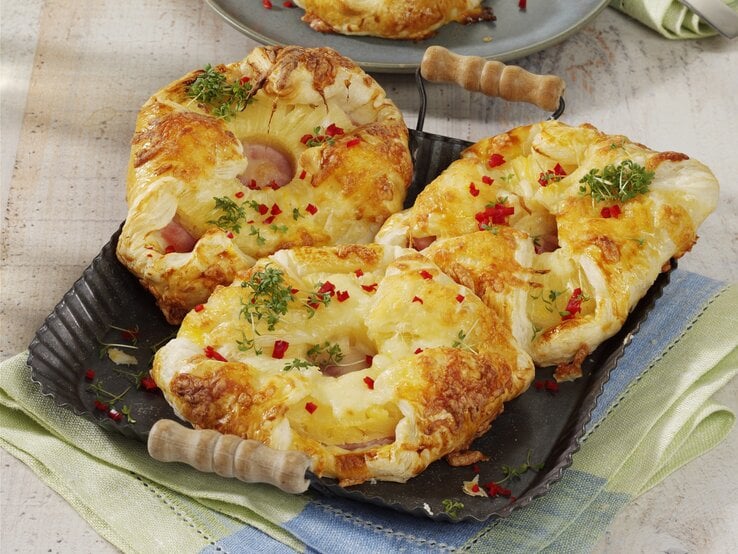 Kopenhagener Teilchen Hawaii, mit Käse überbakcen und mit Petersilie und Chiliflocken garniert, serviert auf einem Tablett auf einem Küchentuch. 