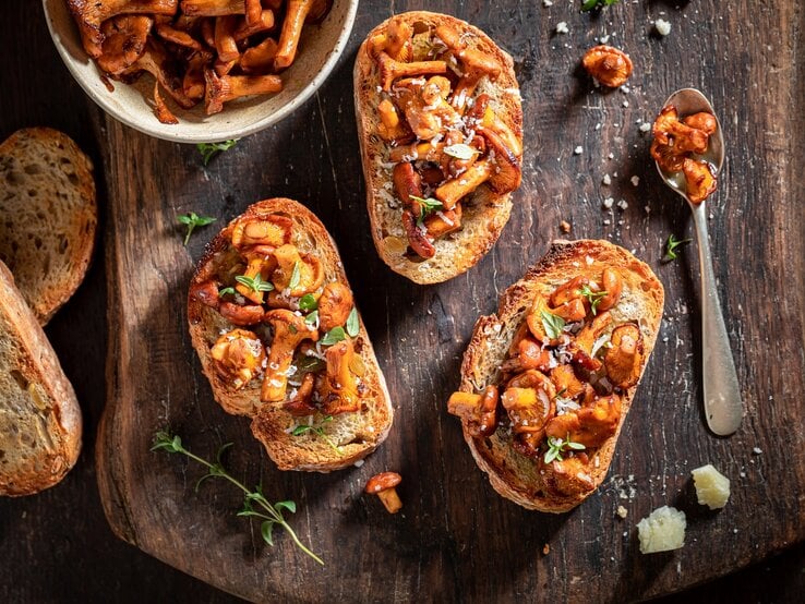Selbstgemachte Knoblauch-Bruschetta mit Pfifferlingen liegen auf einem Untergrund aus Holz. Daneben liegen ein Löffel mit Pilzen, einige Brotscheiben und eine Schüssel mit Pilzen. | © Shutterstock / Shaiith