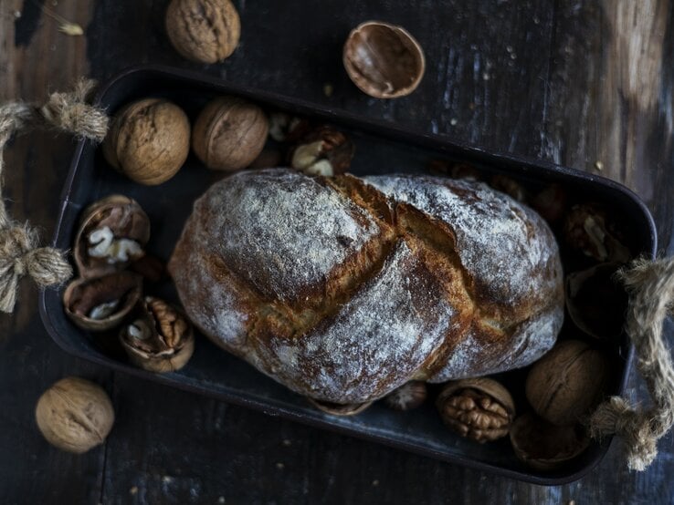 Draufsicht: Auf einem dunklen Untergrund aus Holz liegt ein Walnussbrot in einem dunklen Korb. Drumherum liegen intakte sowie geöffnete Walnüsse.