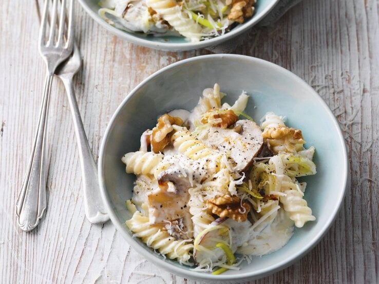 Zwei hellblaue Schalen mit Nudelsalat mit Lauch, Pilzen und Walnüssen steht auf einem Untergrund aus Holz. Daneben liegen zwei Gabeln.
