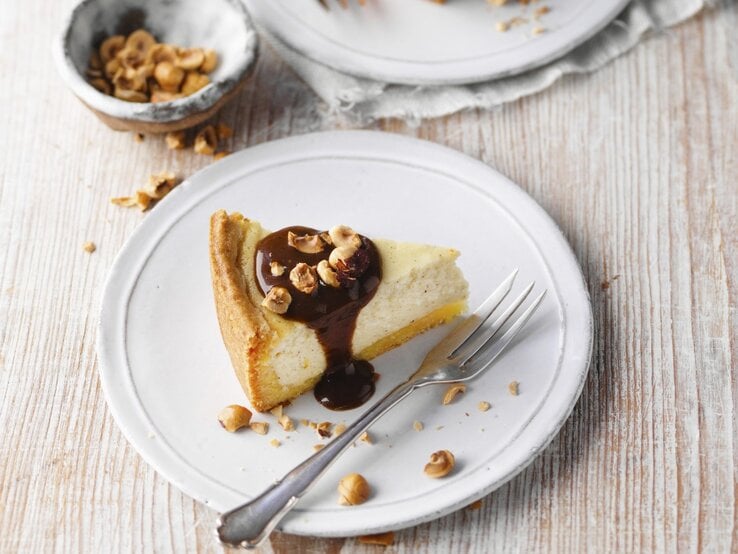 Auf einem Teller ist ein Stück Klatschkiestaat, ein rheinischer Käsekuchen, serviert. Er ist mit Zuckerrübensirup und Haselnüssen getoppt.